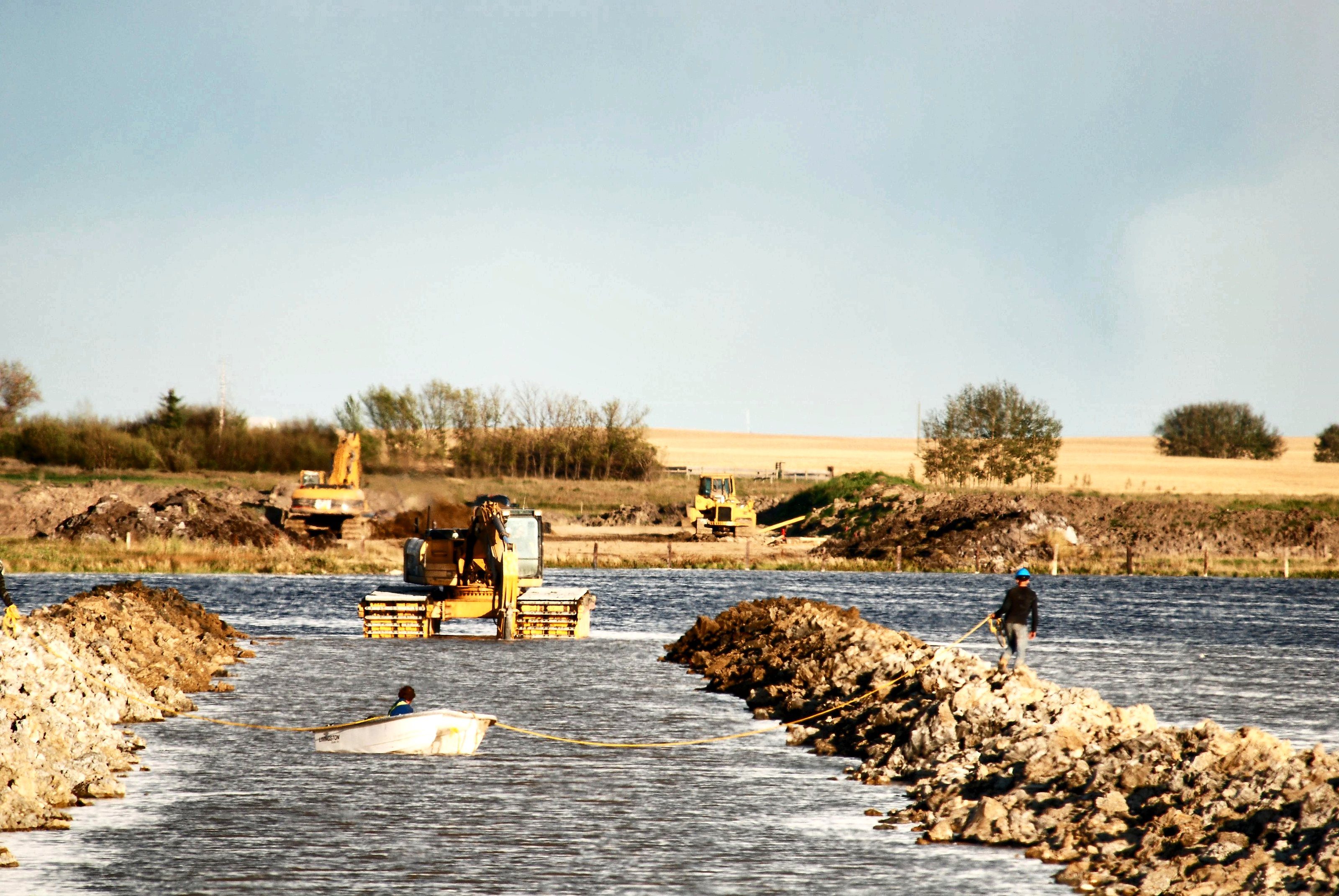 An excavator and a crew working from a rock causeway in shallow water