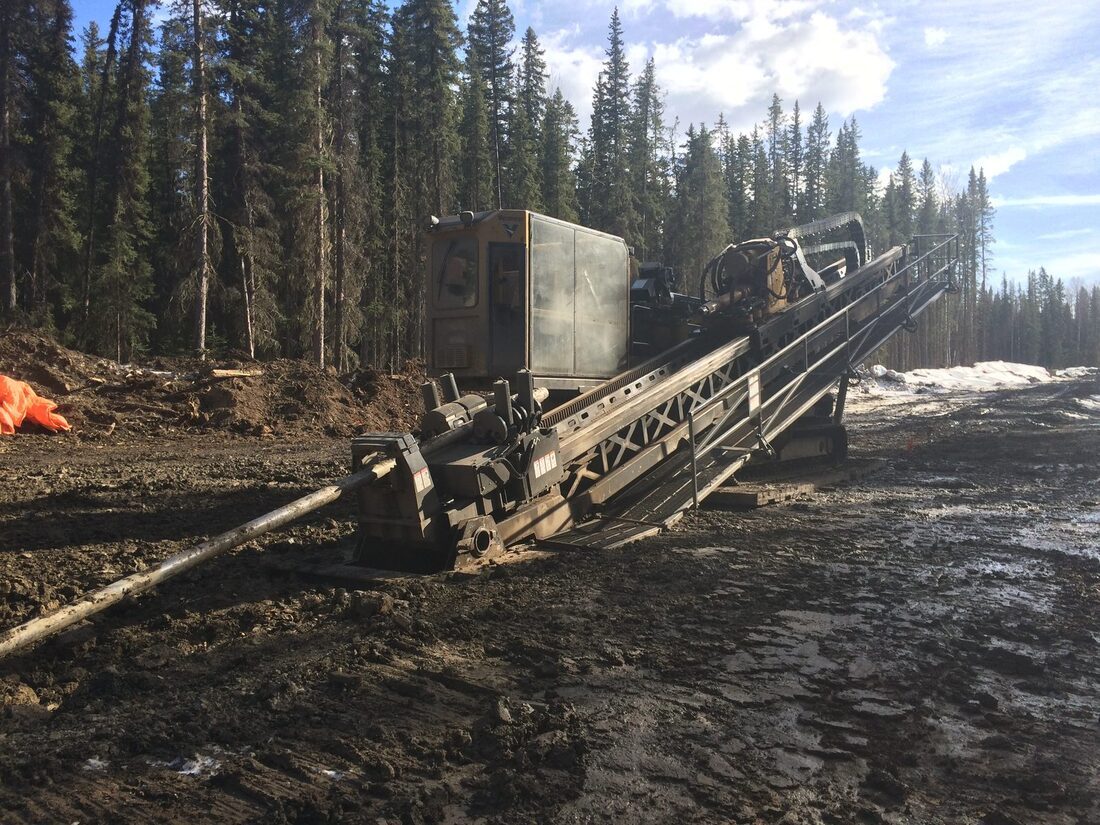 A directional drill rig on tracks with its rod rack laid into the cut, spruce behind