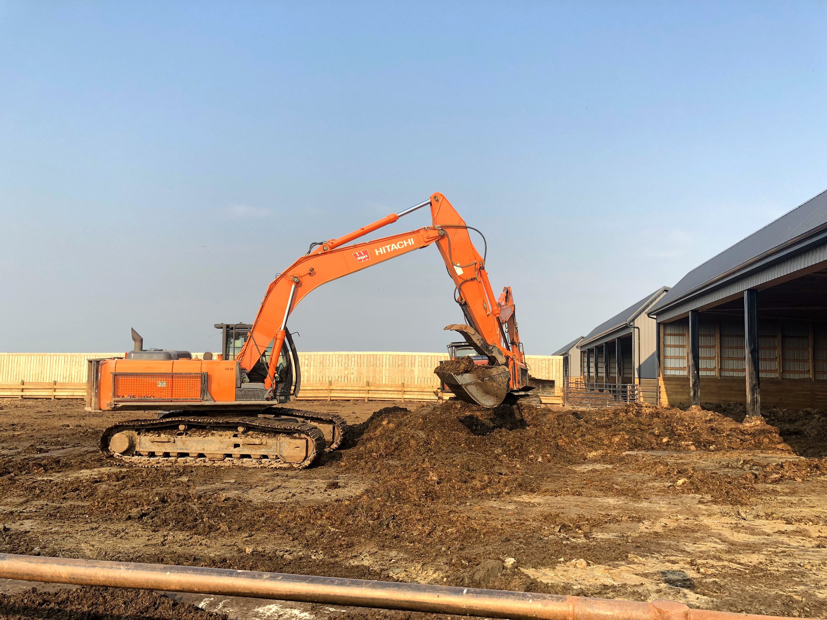 An excavator digging beside a barn, bucket loaded, spoil to the side