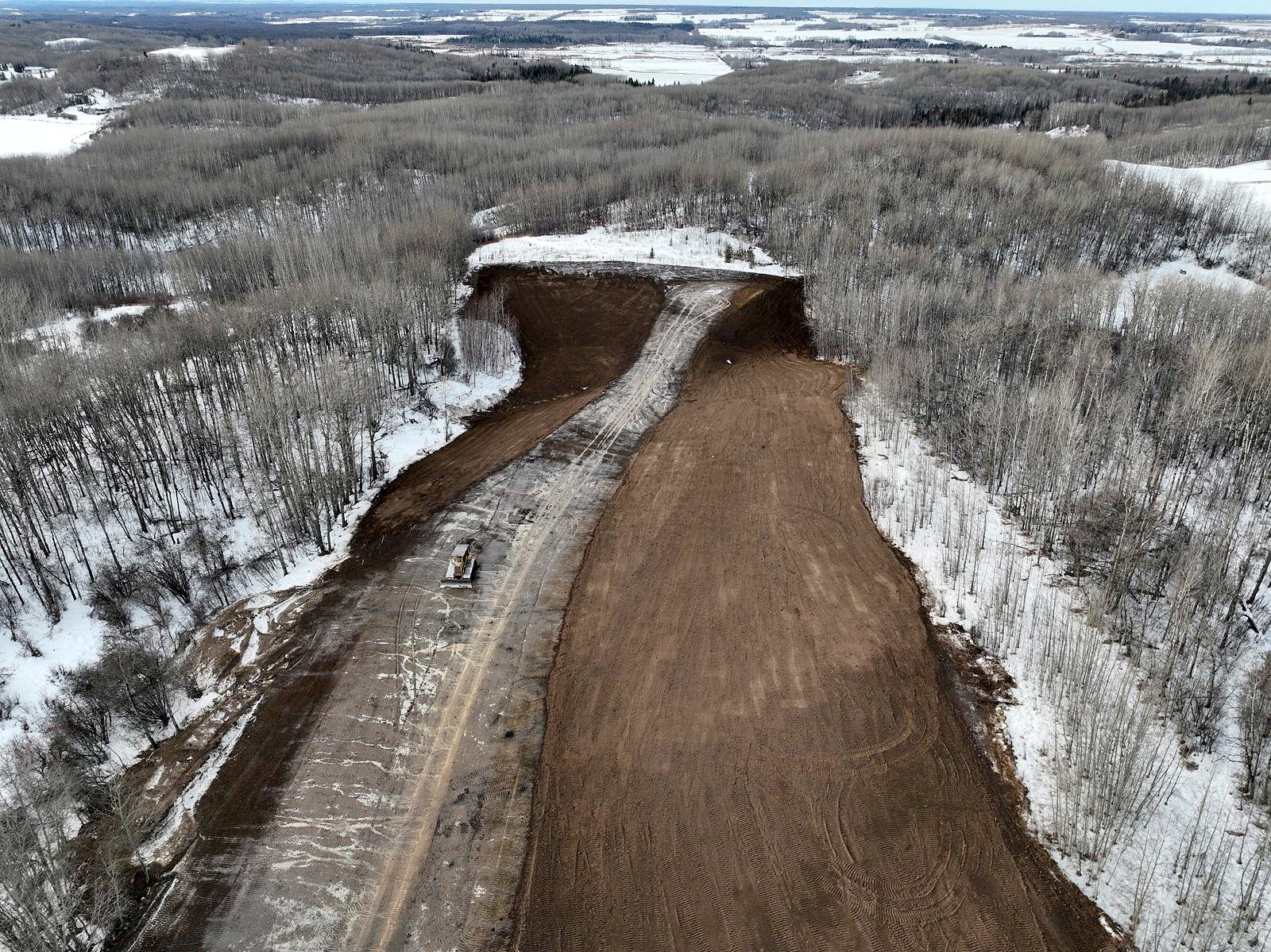 A winter corridor from the air: a graded strip through bush with a dozer working it, snow either side