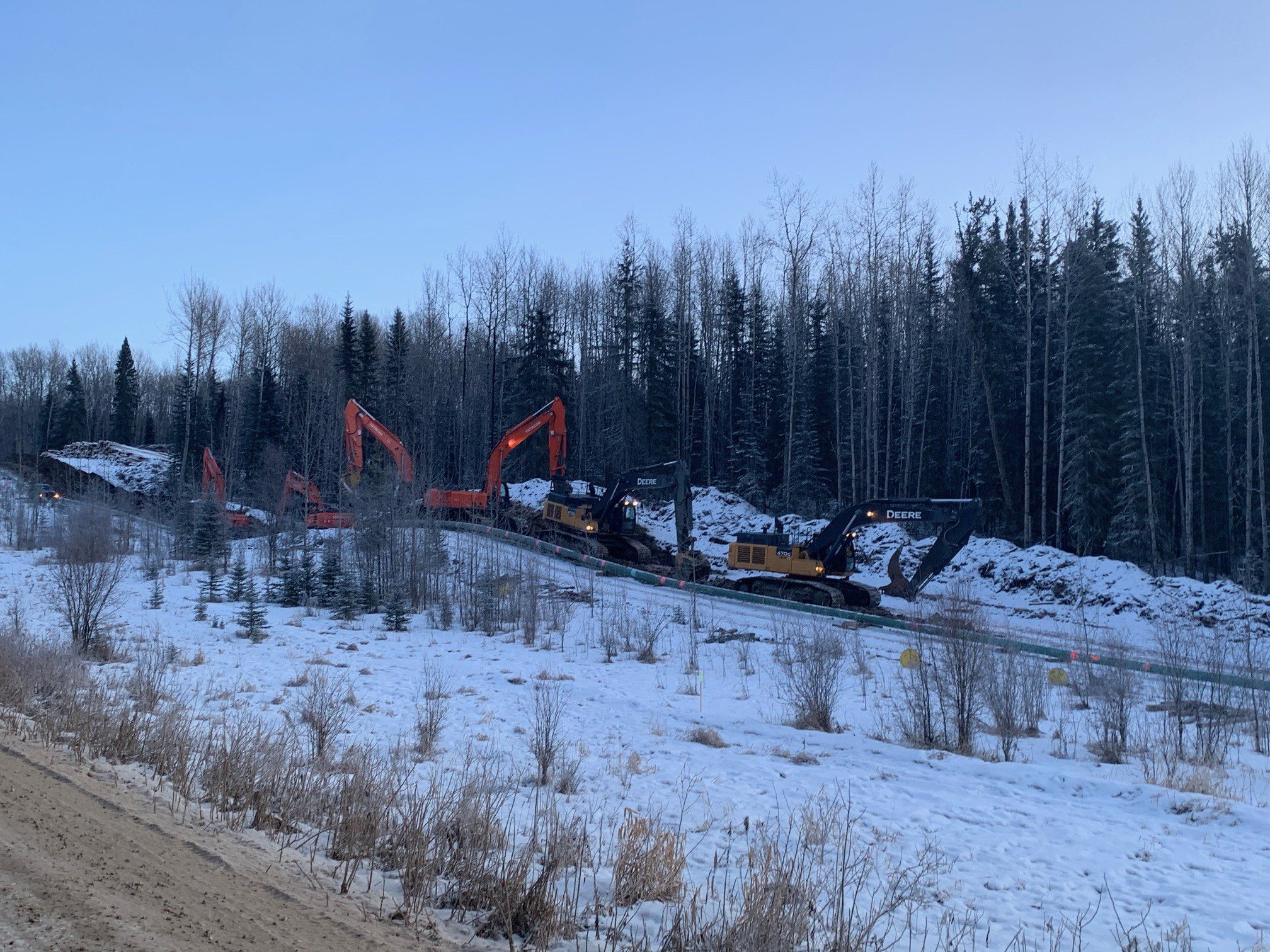 A line of excavators working a cleared corridor in winter, booms up along a strung pipe